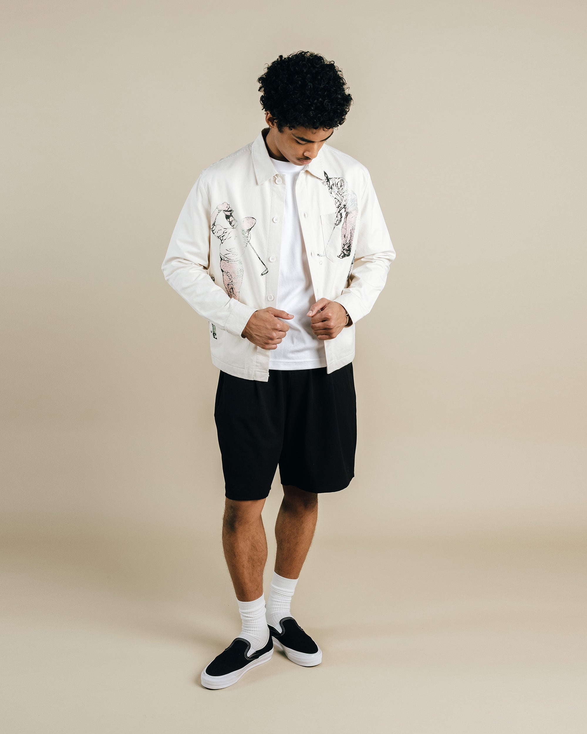Man wearing a white jacket with black patterns, black shorts, and black shoes on a beige background
